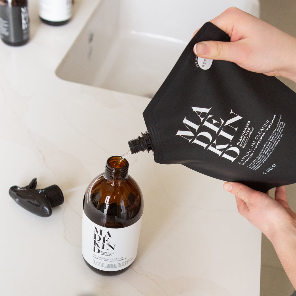 Person pouring liquid from a black 'MadeKind' refill pouch into an amber glass sustainable bottle on a bathroom counter.