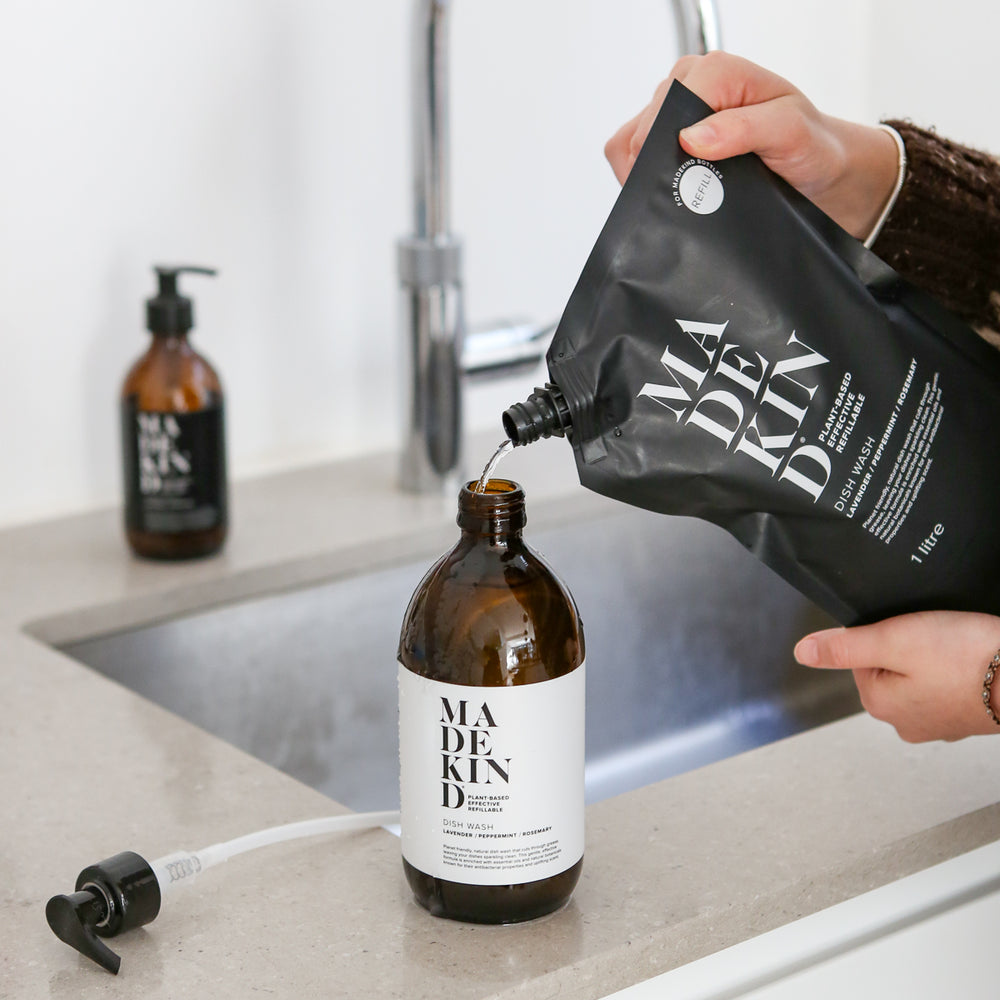 Person pouring liquid from a MadeKind black refill pouch into a refillable glass bottle labeled 'MADEKIND' on a kitchen counter.