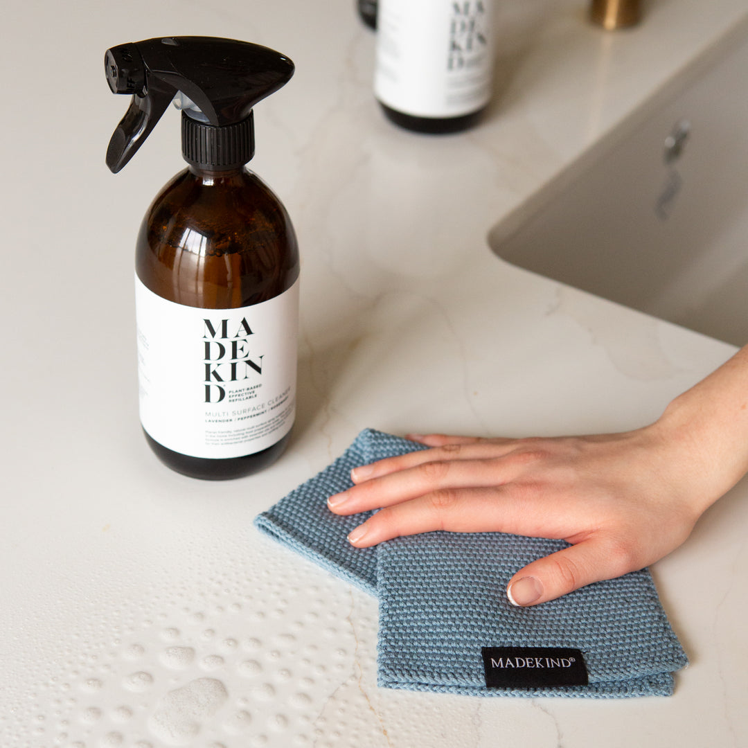 Photo of a hand holding a blue organic cotton dish cloth and a bottle of MadeKind Multi Surface Cleaner on a kitchen counter