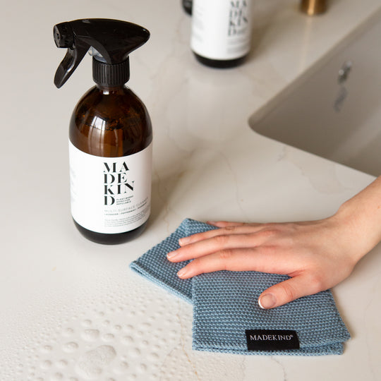 Photo of a hand holding a blue organic cotton dish cloth and a bottle of MadeKind Multi Surface Cleaner on a kitchen counter