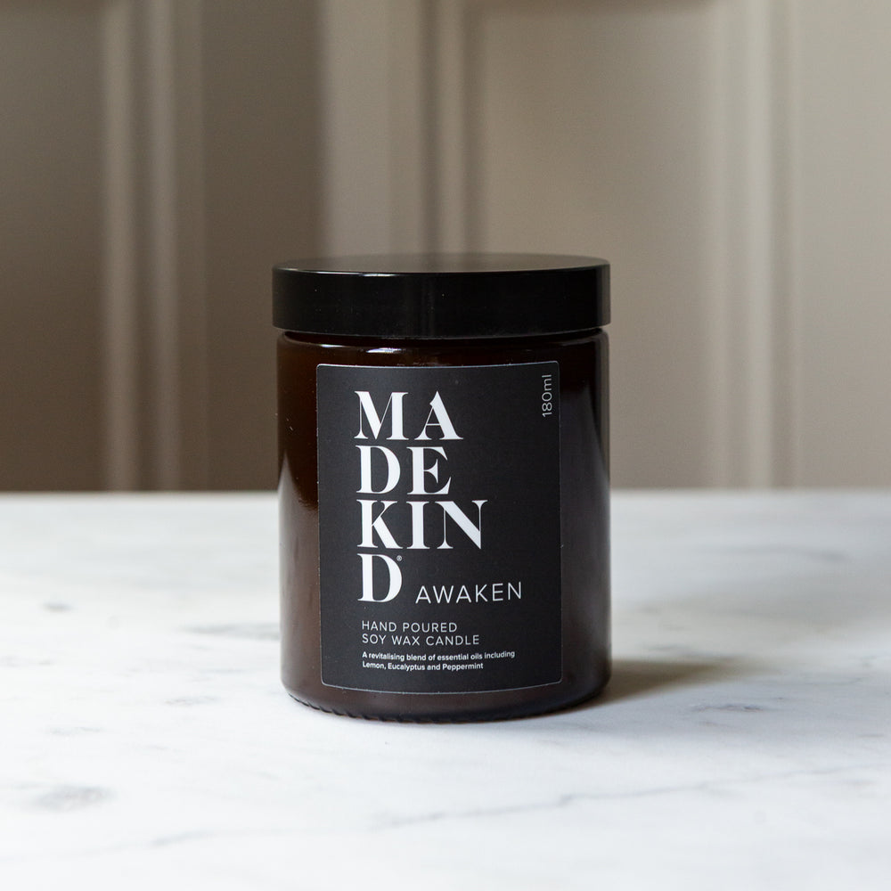 MadeKind Awaken soy wax candle in an amber glass jar candle. Lit with a wooden match held by a person's hand, set on a light marble surface. Scented with essential oils.