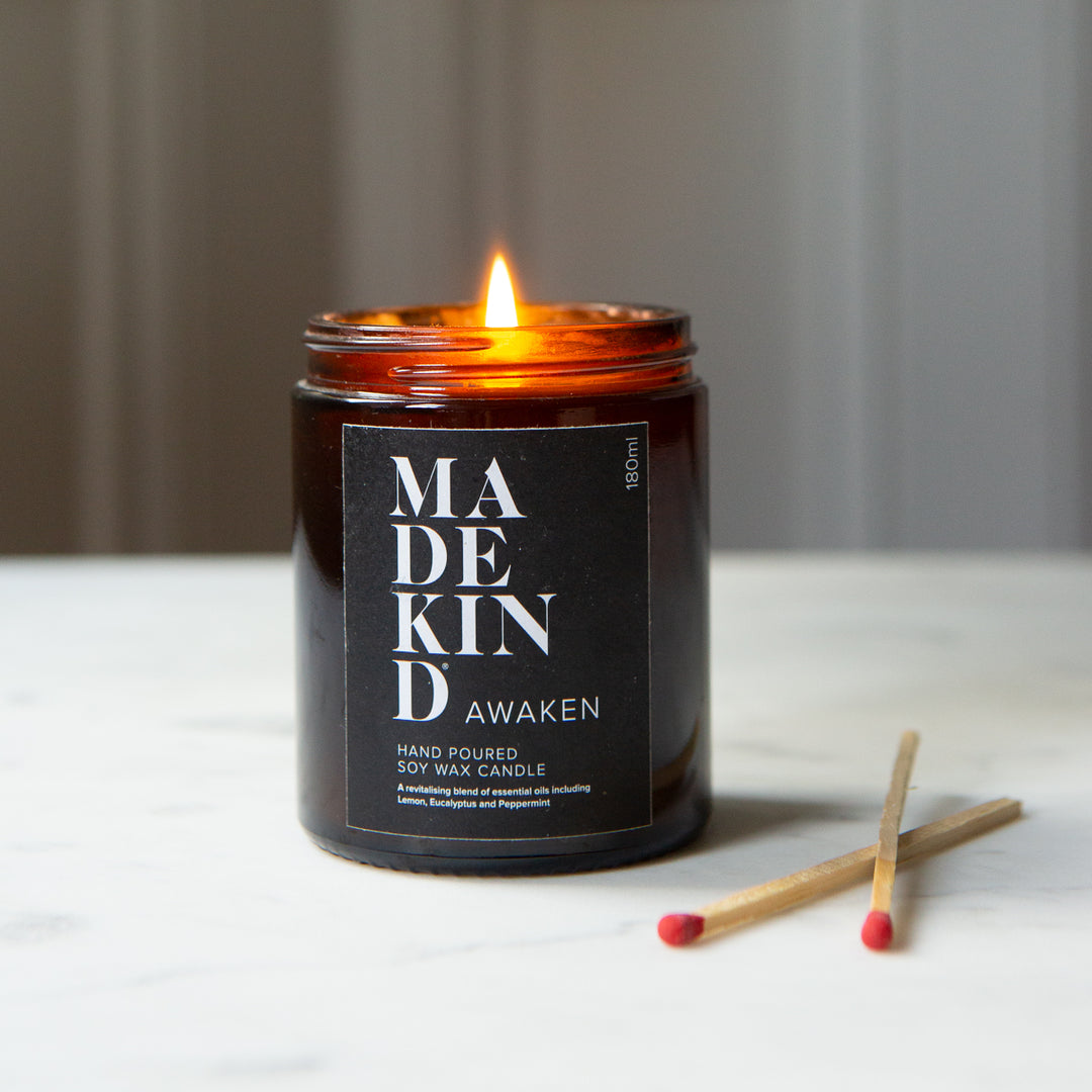 MadeKind Awaken soy wax candle in an amber glass jar candle. Lit with a wooden match held by a person's hand, set on a light marble surface. Scented with essential oils.