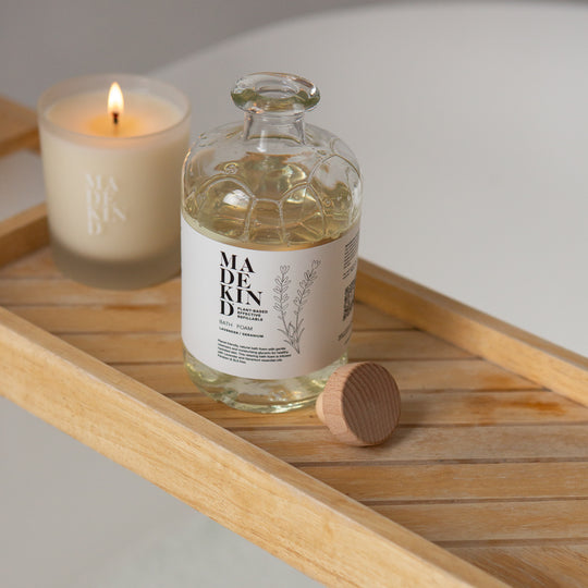 Bottle of MadeKind Bath Foam on a wooden tray with a lit candle in the background over a bath