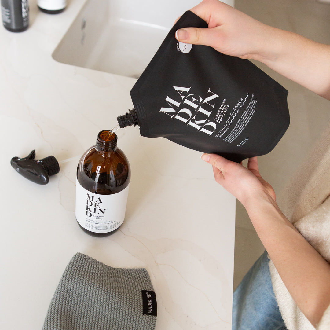 Person pouring Bathroom Cleaner from a black 1 Litre Refill pouch into an amber bottle labeled 'MadeKind' on a bathroom counter.