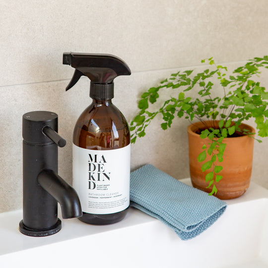 Amber spray bottle with 'MadeKind' label on a sink with a black tap and blue cloth, plant in the background.
