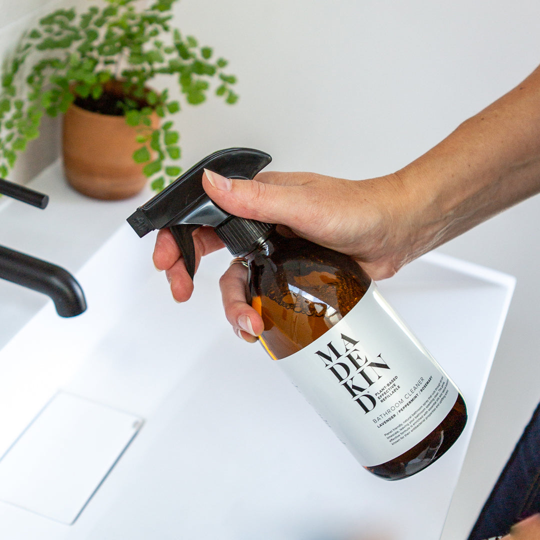 Hand holding a spray bottle labeled 'MadeKind' over a white surface with a plant in the background.