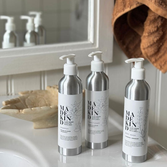 Three aluminium pump bottles of MADEKIND body wash, shampoo and conditioner arranged on a bathroom counter in front of a mirror, with a brown towel hanging on the right.