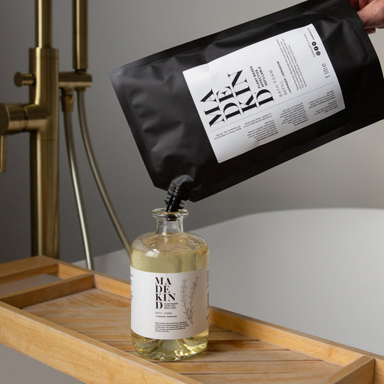 A black refill pouch labeled MadeKind Bath Foam is being tilted to pour a pale yellow liquid into a clear glass bottle with a white label reading MadeKind Bath Foam, set on a wooden tray with a brass shower fixture in the background.