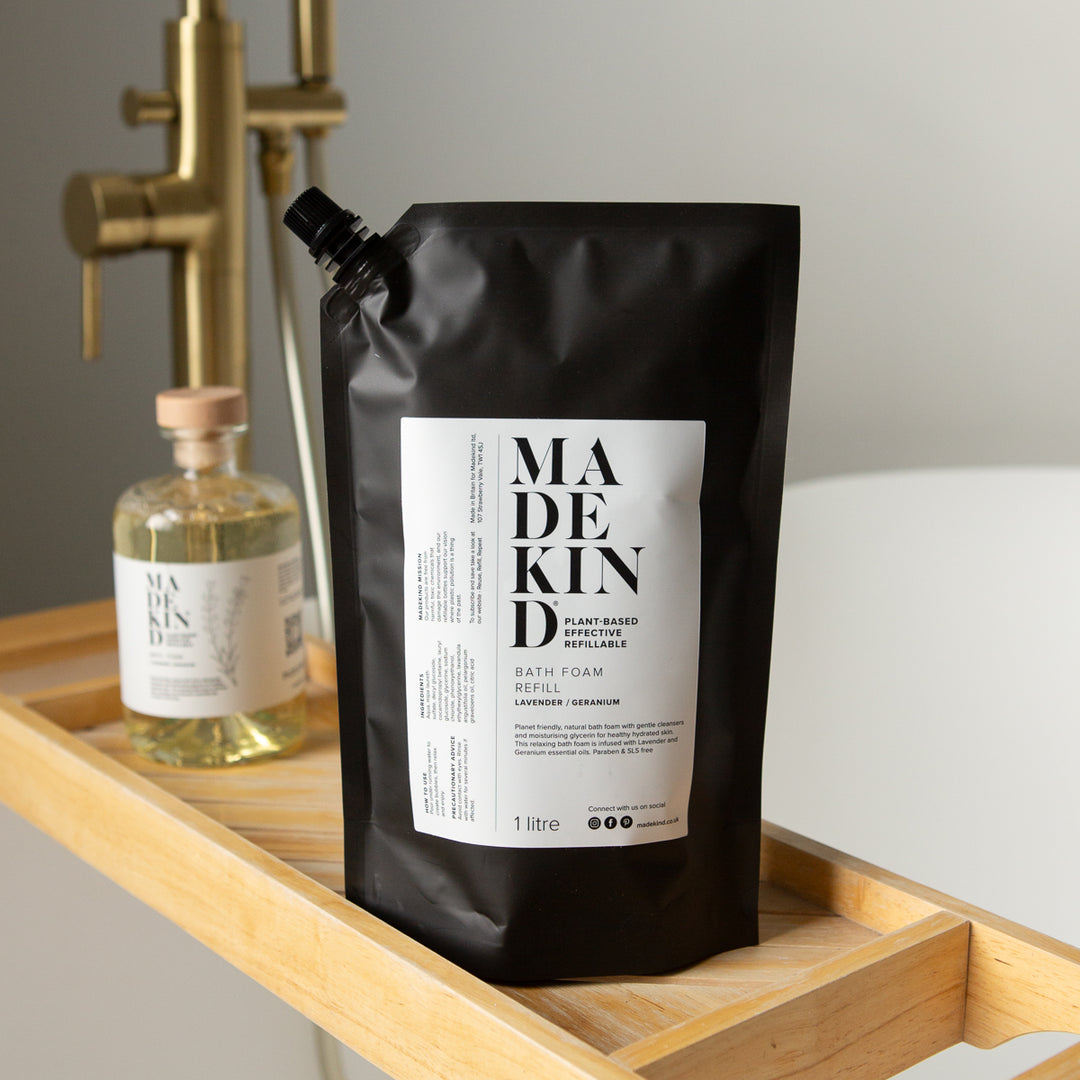 A black stand-up pouch of MadeKind bath foam sits on a wooden bath tray beside a MadeKind Bath Foam glass bottle, with a brass shower fixture and white bathtub in the background.