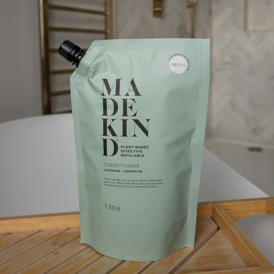 A refillable conditioner pouch by MadeKind in a pale mint green colour, standing on a wooden tray in front of a bath.