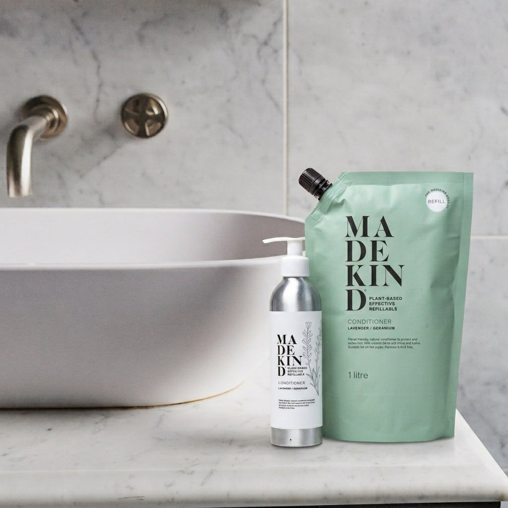 MadeKind natural haircare products shown on a bathroom counter: a silver pump bottle and a mint green 1L refill pouch beside a white sink in a marble bathroom.