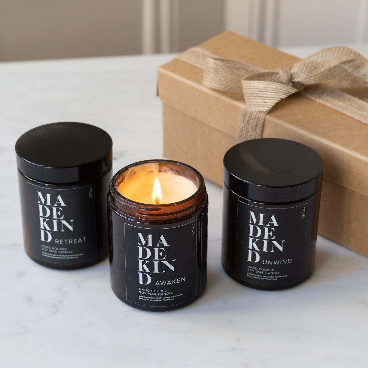 Three MadeKind soy wax candles candles in amber glass jars with black lids are arranged on a light marble surface, with one candle lit in the centre. A kraft gift box with a burlap ribbon sits behind them.
