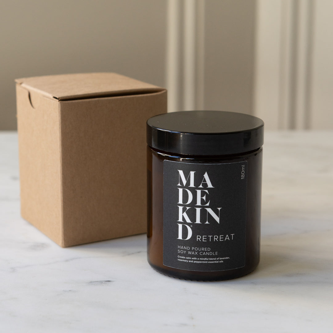 MadeKind soy wax candle in an amber glass jar candle. Lit with a wooden match held by a person's hand, set on a light marble surface. Scented with essential oils to make your home smell like a retreat.