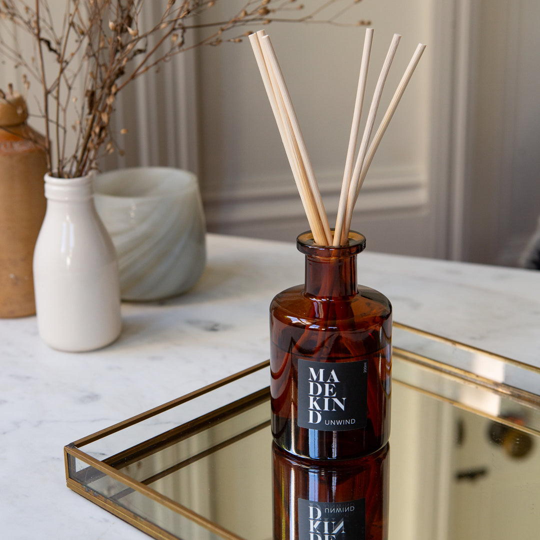 Amber glass reed diffuser bottle with natural wood reeds on a mirrored tray, set on a white marble table with decorative vases in the background. The bottle label reads 'MADEKIND'.