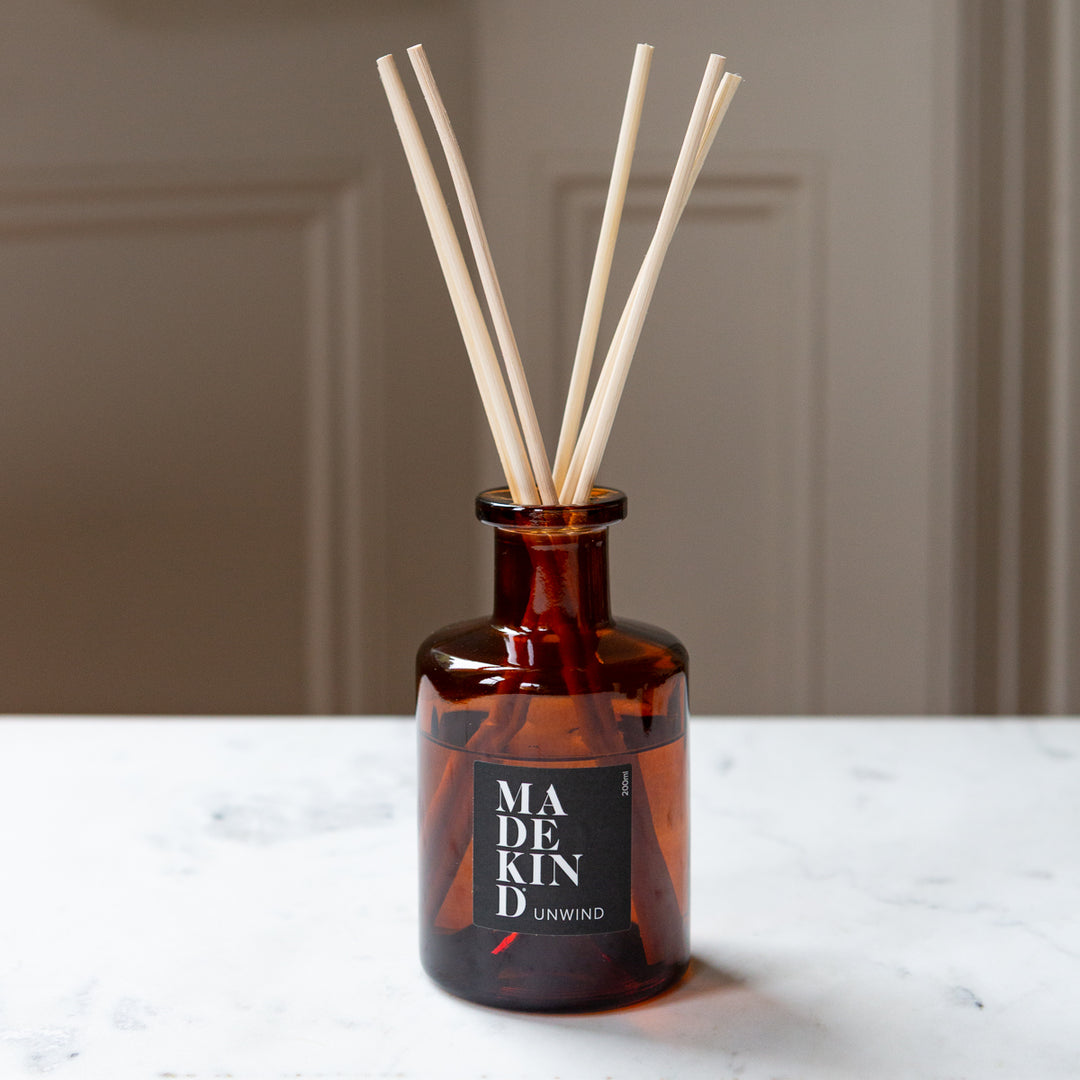 Amber glass reed diffuser bottle with six natural reed sticks, featuring a black label that reads 'MADE KIND UNWIND', set on a white marble surface against a neutral beige background.