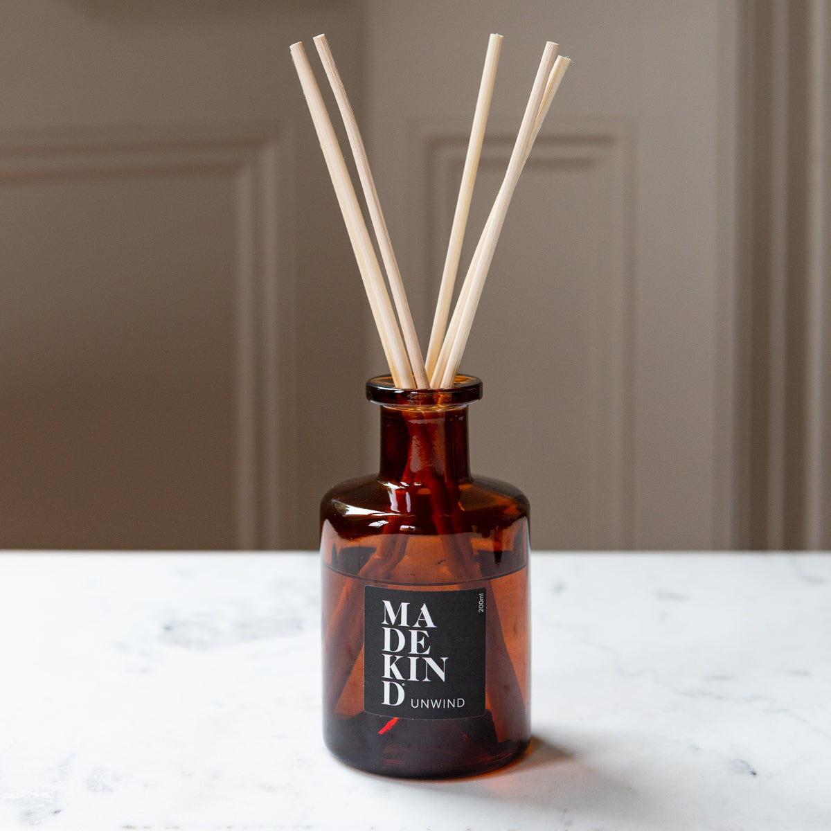 Amber glass reed diffuser bottle with six natural reed sticks, featuring a black label that reads 'MADE KIND UNWIND', set on a white marble surface against a neutral beige background.