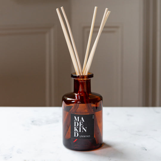 Amber glass reed diffuser bottle with six natural reed sticks, featuring a black label that reads 'MADE KIND UNWIND', set on a white marble surface against a neutral beige background.
