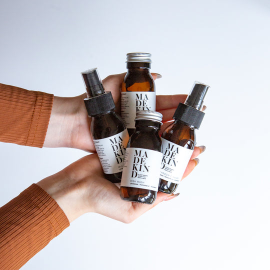 A set of MadeKind mini cleaning products being held in hands. The set includes four amber glass bottles with cleaning products in, with white labels and metal caps, plus a cream canvas zip pouch with black text.