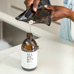 Photo of lady using 1 litre multi surface cleaner refill pouch to fill 500ml amber glass bottle on kitchen counter