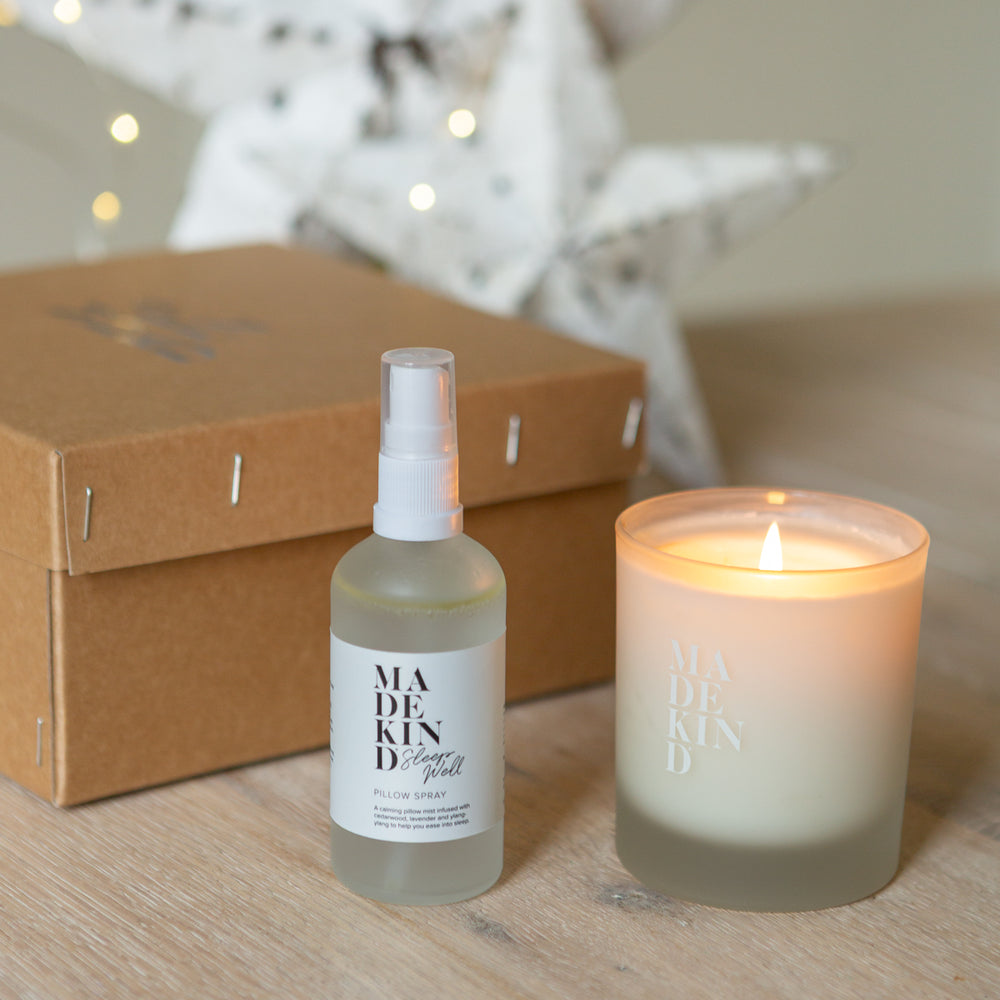 Madekind pillow mist and soy wax aromatherapy sleep well candle on a wooden surface next to a gift box with a blurred starry background