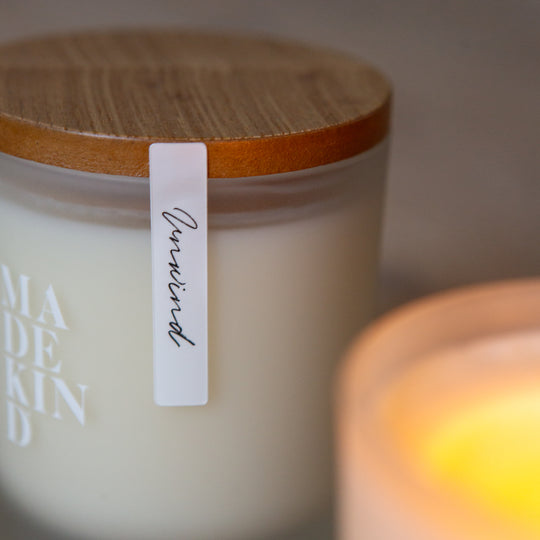 Two candles, one lit and one unlit, on a wooden surface with wooden lid with 'MADEKIND' branding, in unwind blend for relaxation