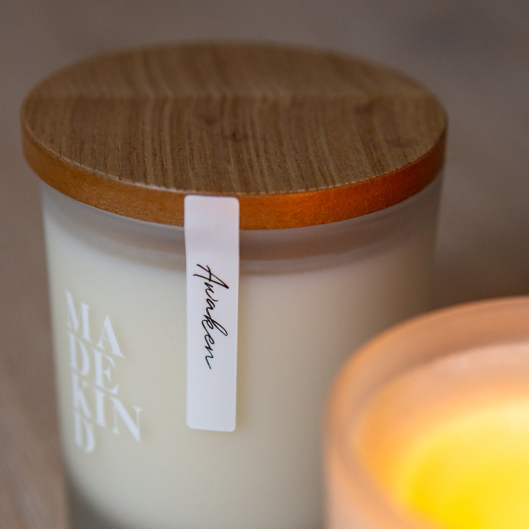 Frosted glass candles on a wooden surface, one lit and labeled 'MADE KIND'. Soy wax candle with wood lid.