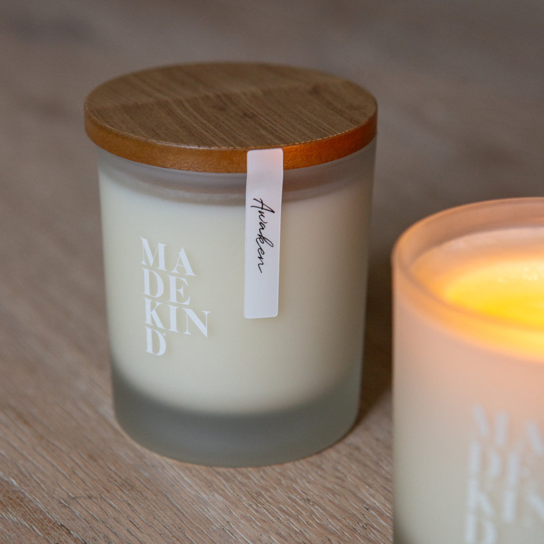 Frosted glass candles on a wooden surface, one lit and labeled 'MADE KIND'. Soy wax candle with wood lid.