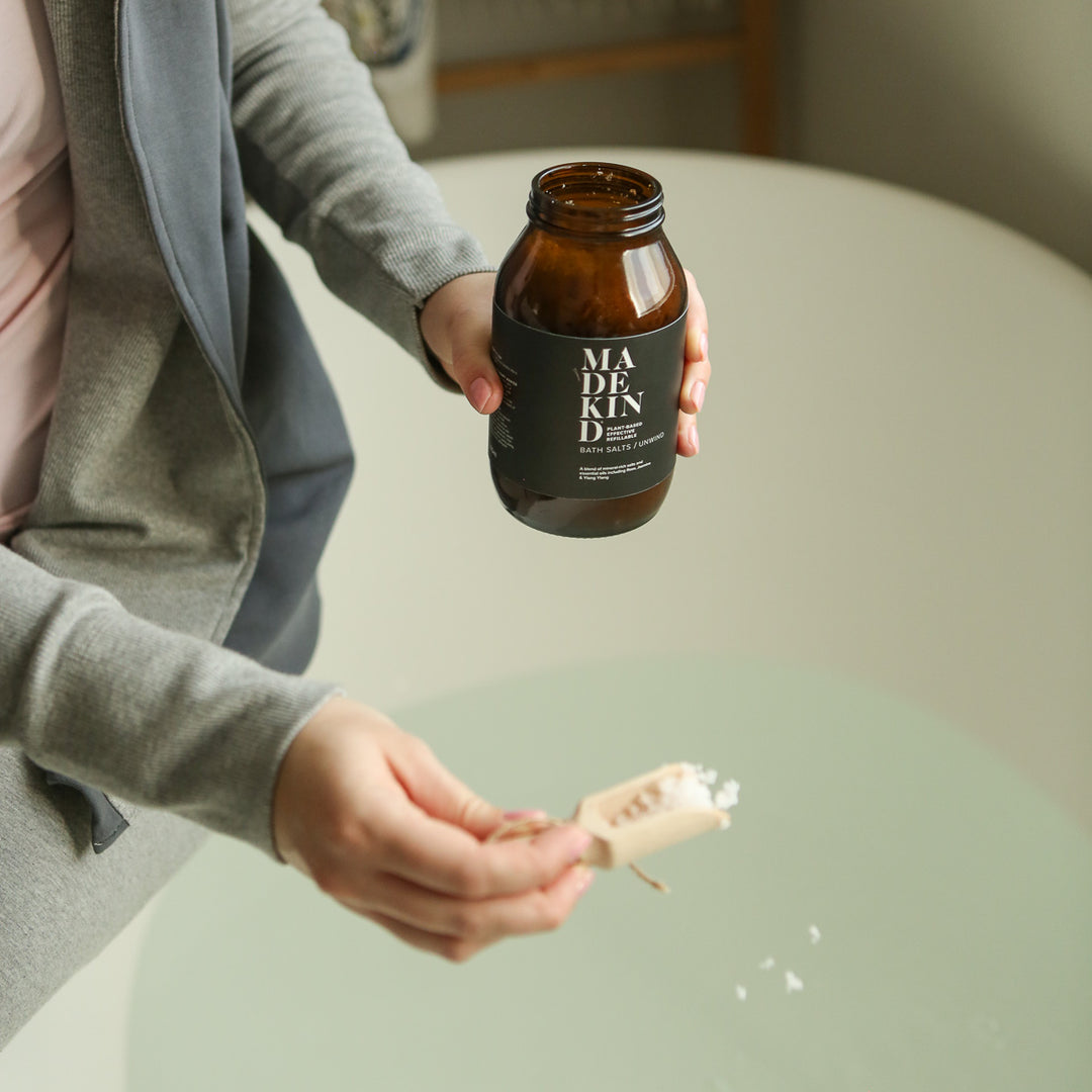 Photo of a person holding an amber glass jar full of bath salts whilst they are holding a scoop full of bath salts over a bath