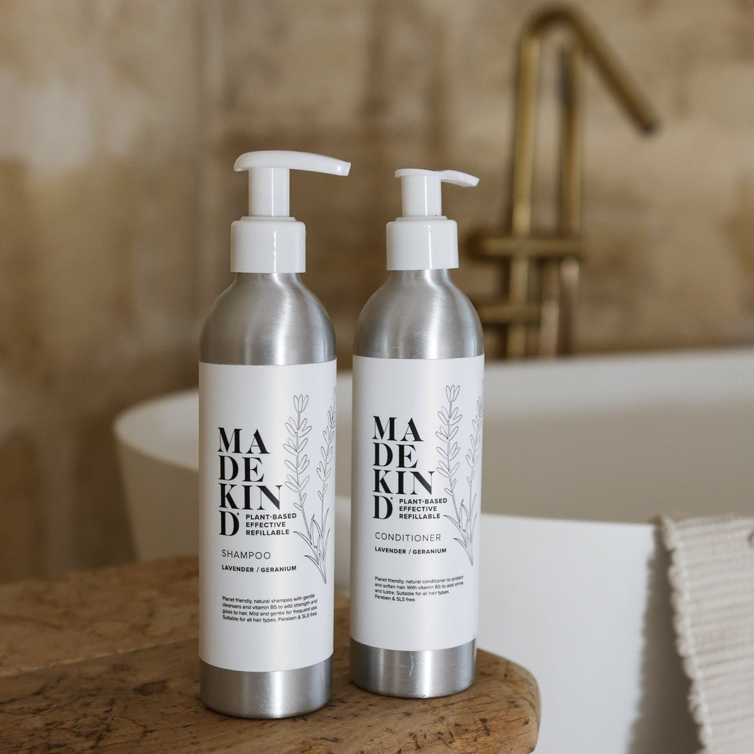 Two silver pump bottles of MadeKind natural shampoo and conditioner with white labels, standing on a wooden block next to a white sink in a bathroom setting.