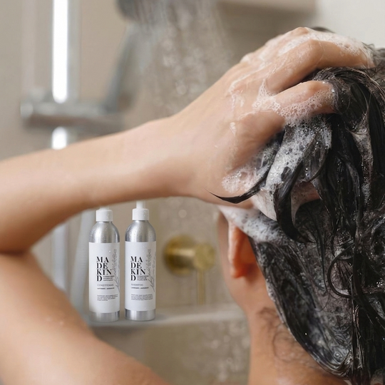 A person washing their hair in a shower, with eco-friendly natural shampoo lather on dark hair. Two silver bottles of MadeKind shampoo and conditioner are shown in the lower left foreground against a softly blurred bathroom background.