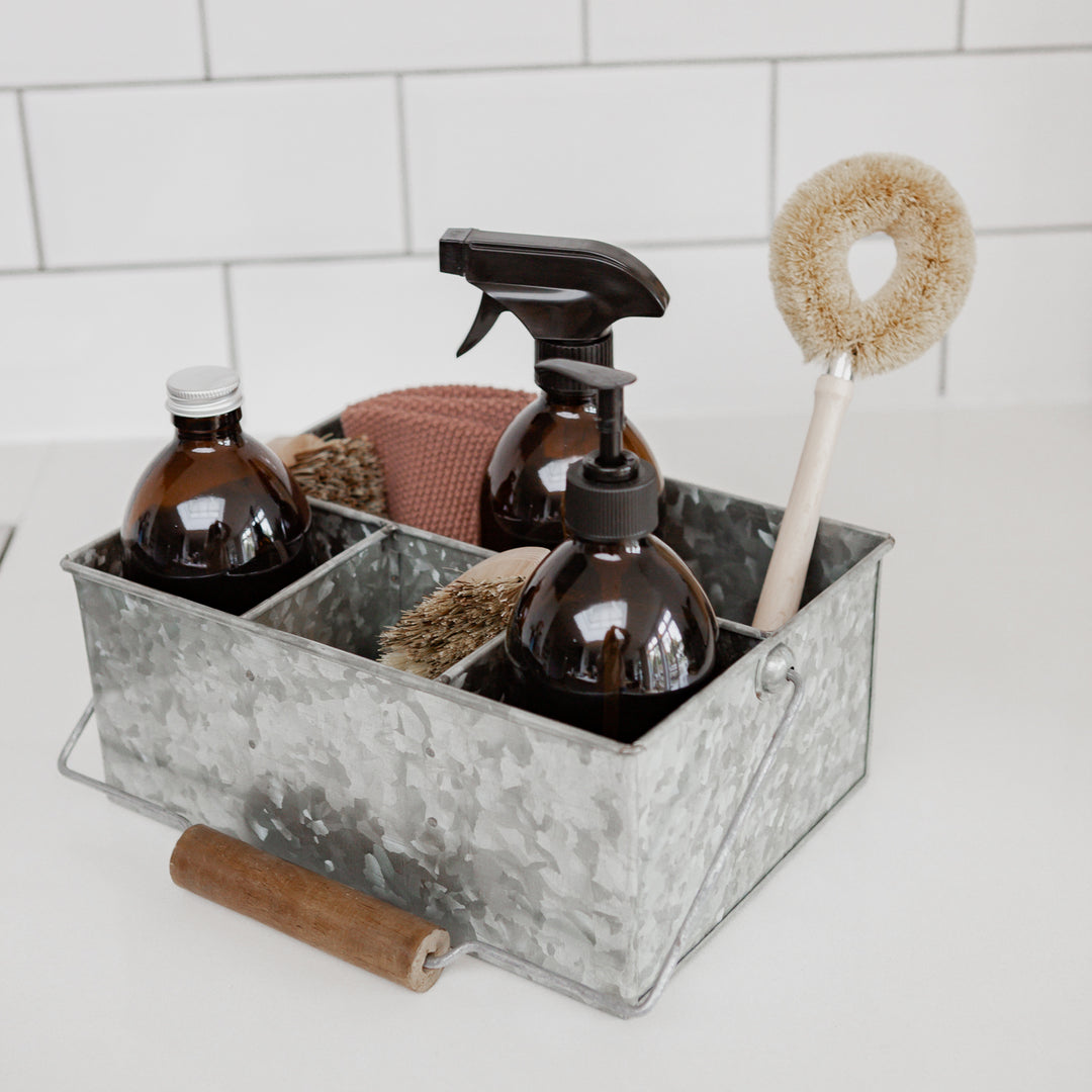 Photo of three brushes, a cloth and 3 three amber glass bottles sitting in a metal cleaning caddy with a white brick backdrop