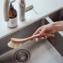 Load image into Gallery viewer, Photo of a Redecker curved handle wooden dish brush being held over a kitchen sink
